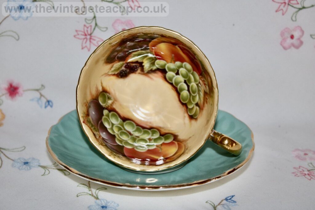 This exquisite set of three Aynsley bone china teacup trios features the iconic Orchard Gold design, a highly collectible and sought-after pattern known for its vibrant hand-painted fruit motif set inside a gold-gilded interior. Each trio includes a teacup, saucer, and side plate, all finished in a rich turquoise blue with elegant gold trim on the rims and handles. The interiors of the teacups are ornately painted with peaches, grapes, and leaves, all set against a lustrous gold background.